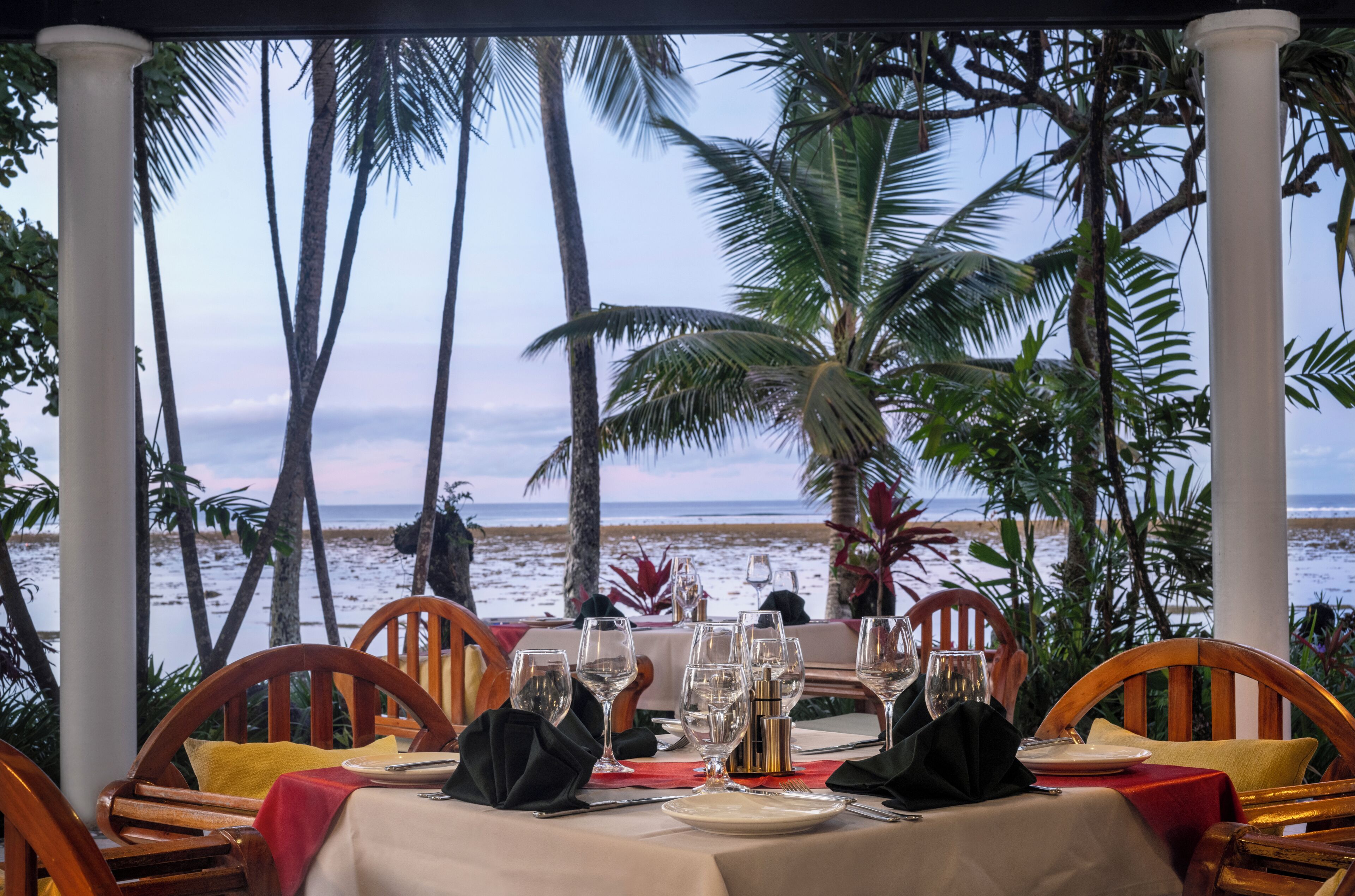 A table set for dining with a view of the beach and palm trees.