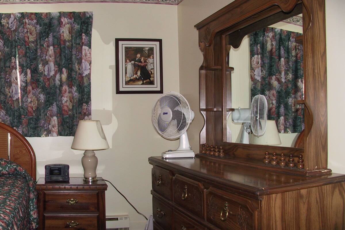 Quarto com cama de madeira, mesa de cabeceira com abajur e ventilador, cômoda e duas fotos emolduradas na parede.