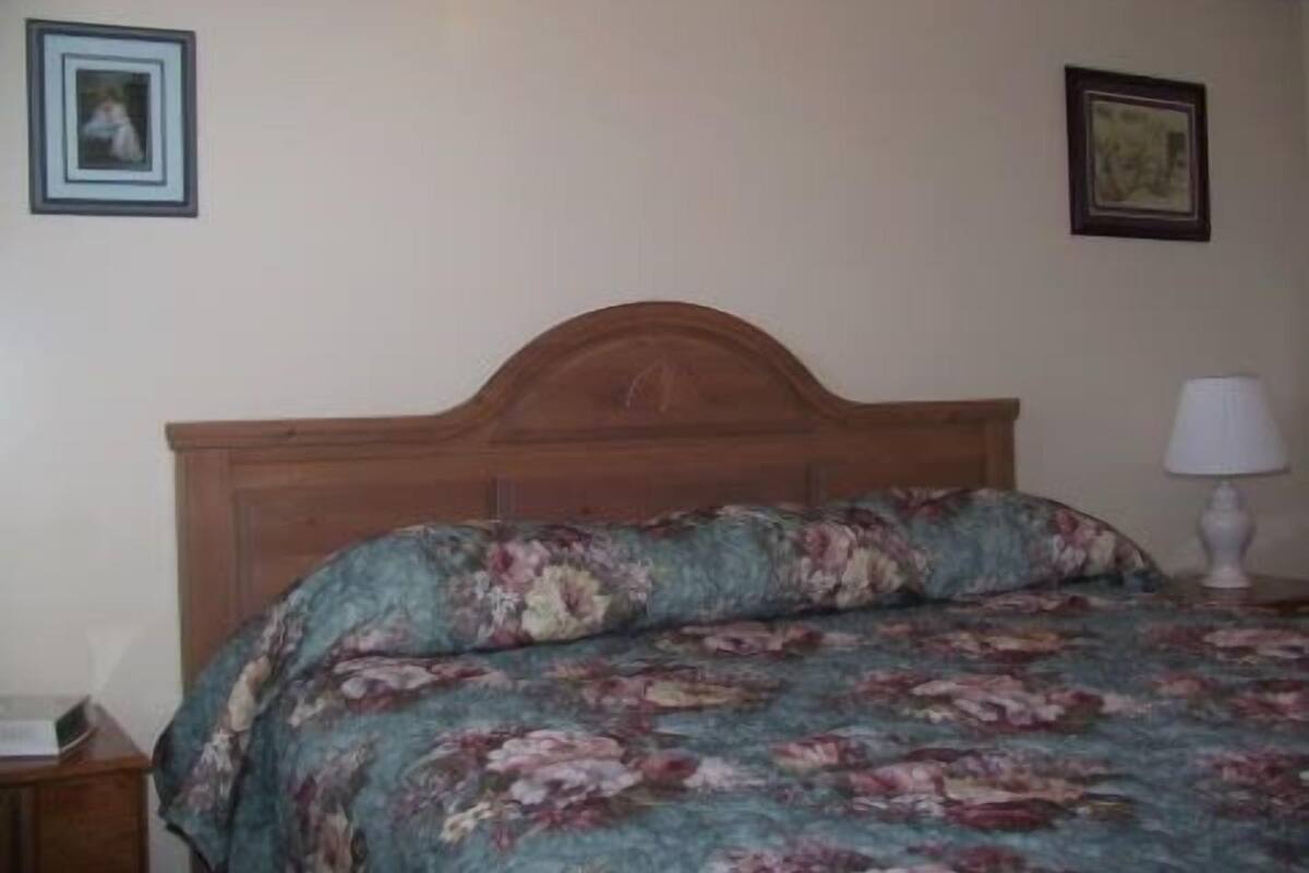 Quarto com cabeceira de madeira, roupa de cama floral, mesa de cabeceira com abajur e duas imagens emolduradas na parede.