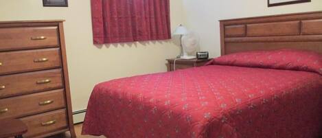 A bedroom with a wooden dresser, a bed with a red bedspread, a nightstand with a lamp, and a framed picture on the wall.