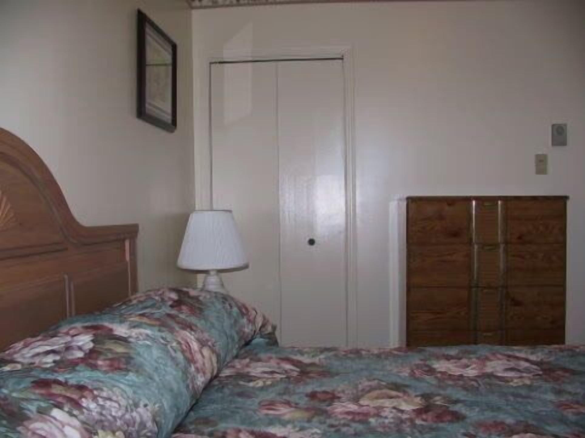 Quarto com cama de madeira, colcha florida, abajur e guarda-roupa.
