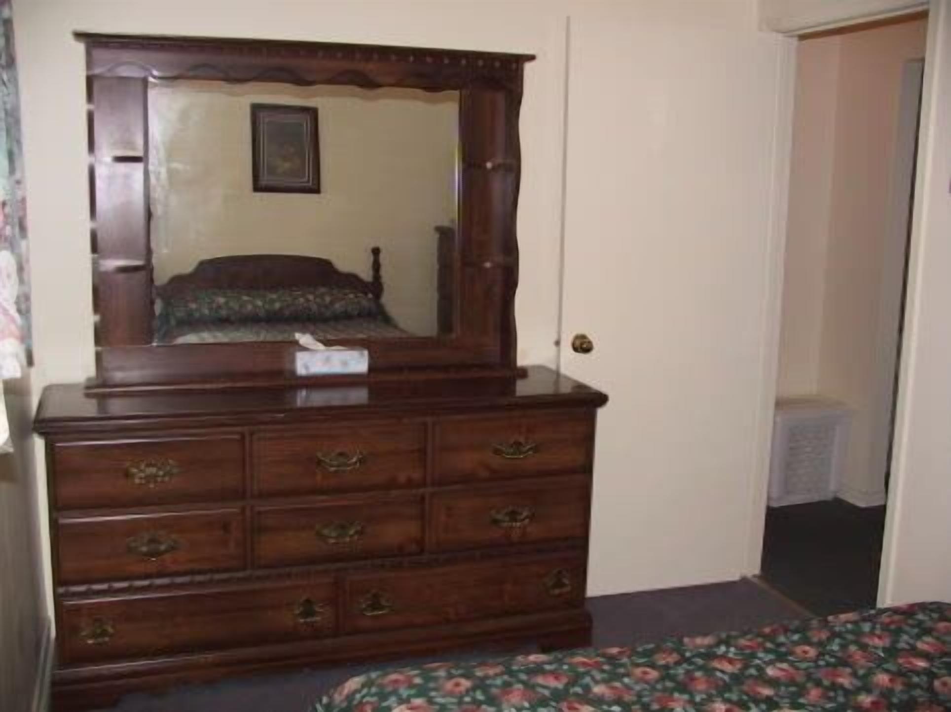 A wooden dresser with multiple drawers and a mirror, a bed with floral bedding, and a door leading to another room.