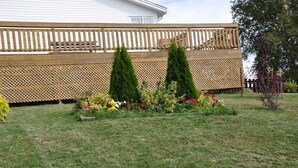 A fenced backyard with a wooden deck, two coniferous trees, and a variety of colorful flowers.