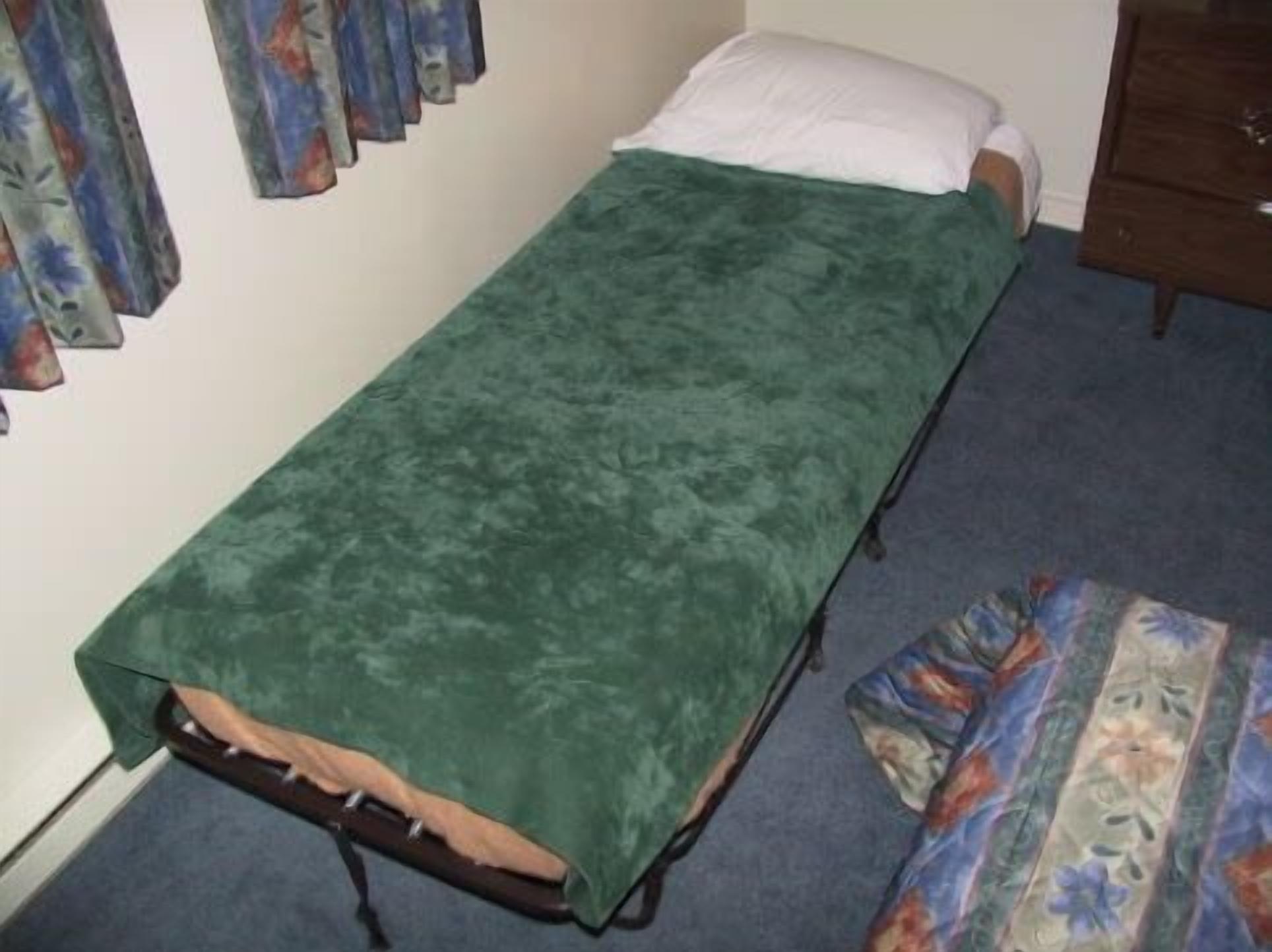 A small single bed with a green bedspread, a wooden headboard, and a floral-patterned rug on the floor.