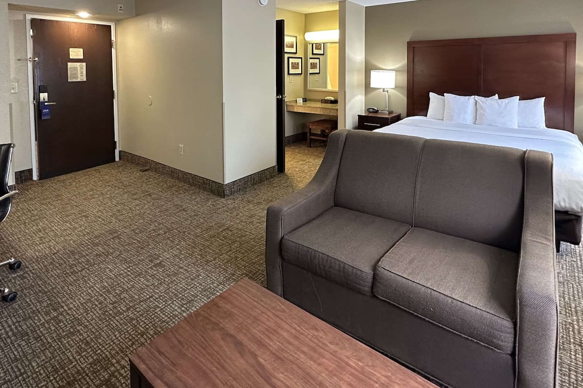 A hotel room with a bed, a sofa, a small table, a desk, a chair, a bathroom, and a door.