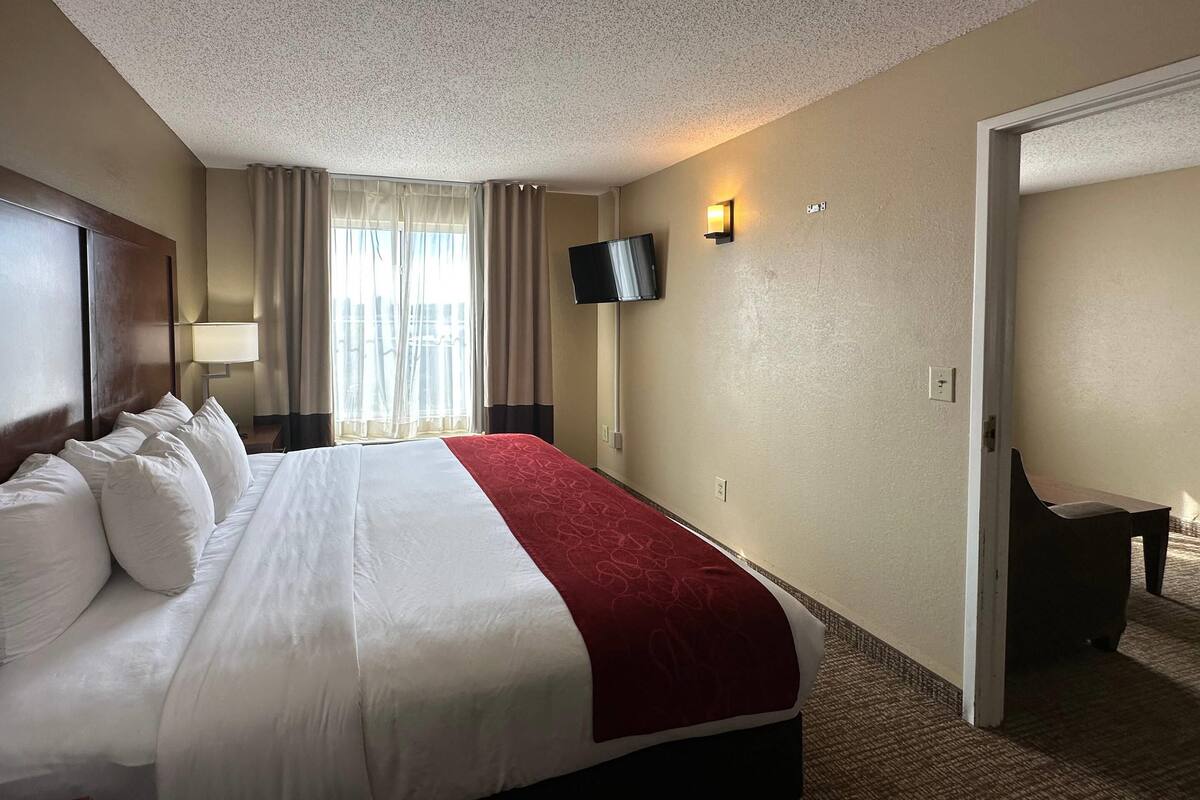 A hotel room with a large bed, a TV mounted on the wall, a desk with a chair, and a door leading to another room.
