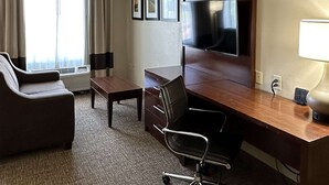 A hotel room with a sofa, a desk with a chair, a flat-screen TV, a lamp, and a window with curtains. - Charlotte Suites (Charlotte)