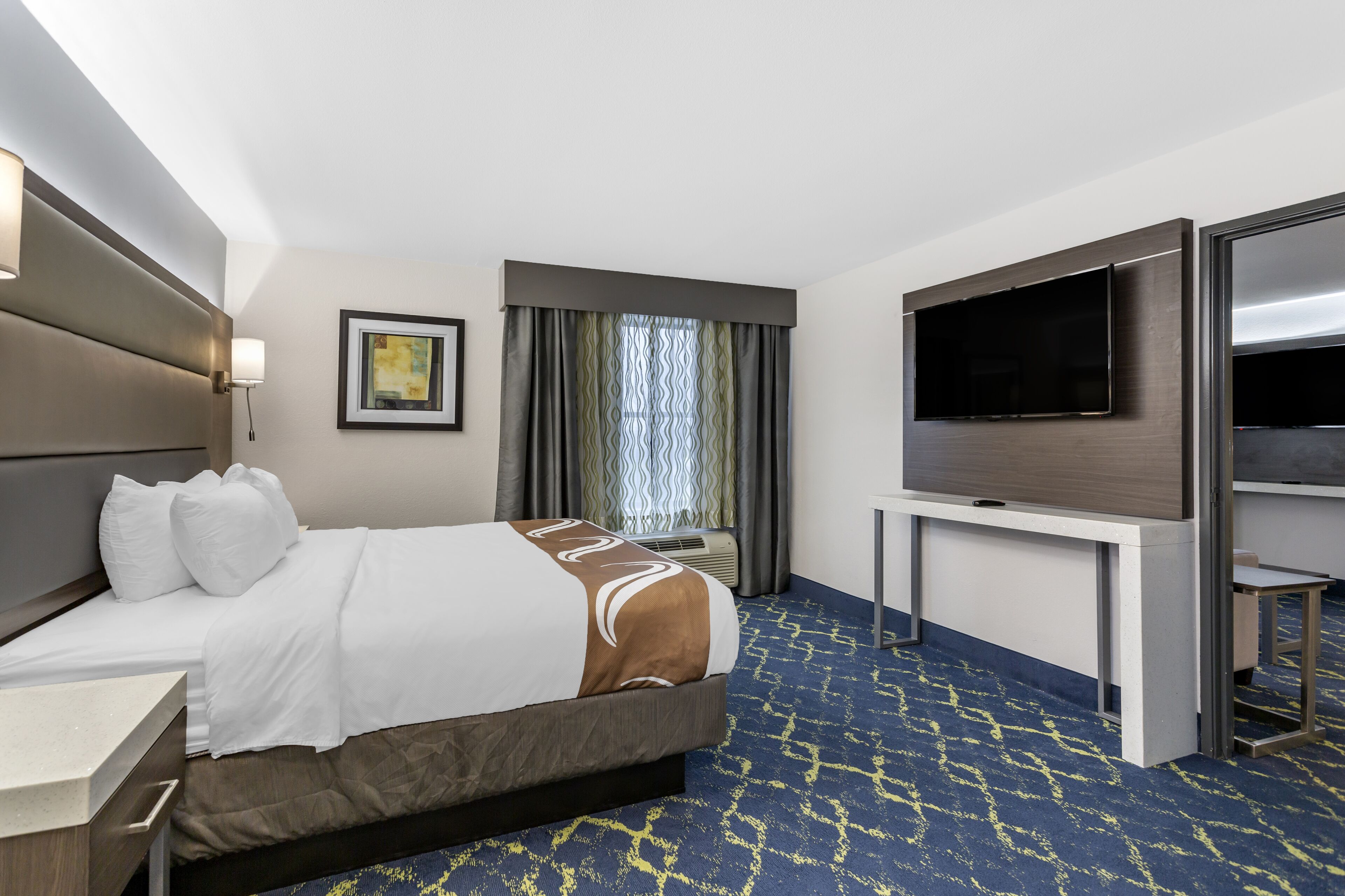 A hotel room with a large bed, a flat-screen TV, a desk, and a window with curtains.
