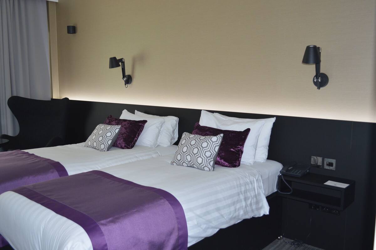 Habitación de hotel con una cama grande, dos mesitas de noche, un teléfono y una lámpara en la pared.