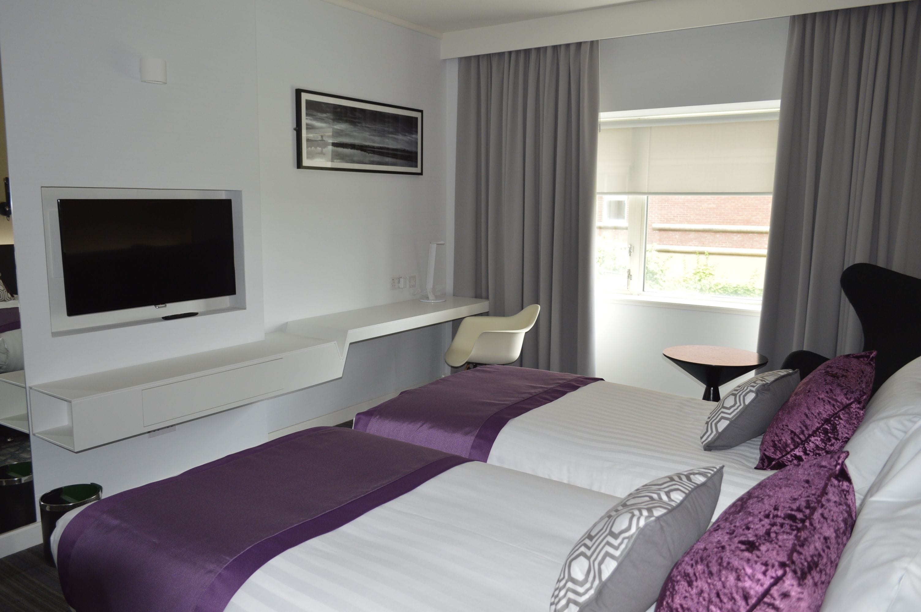 A hotel room with two beds, a TV, a desk, and a window with curtains.