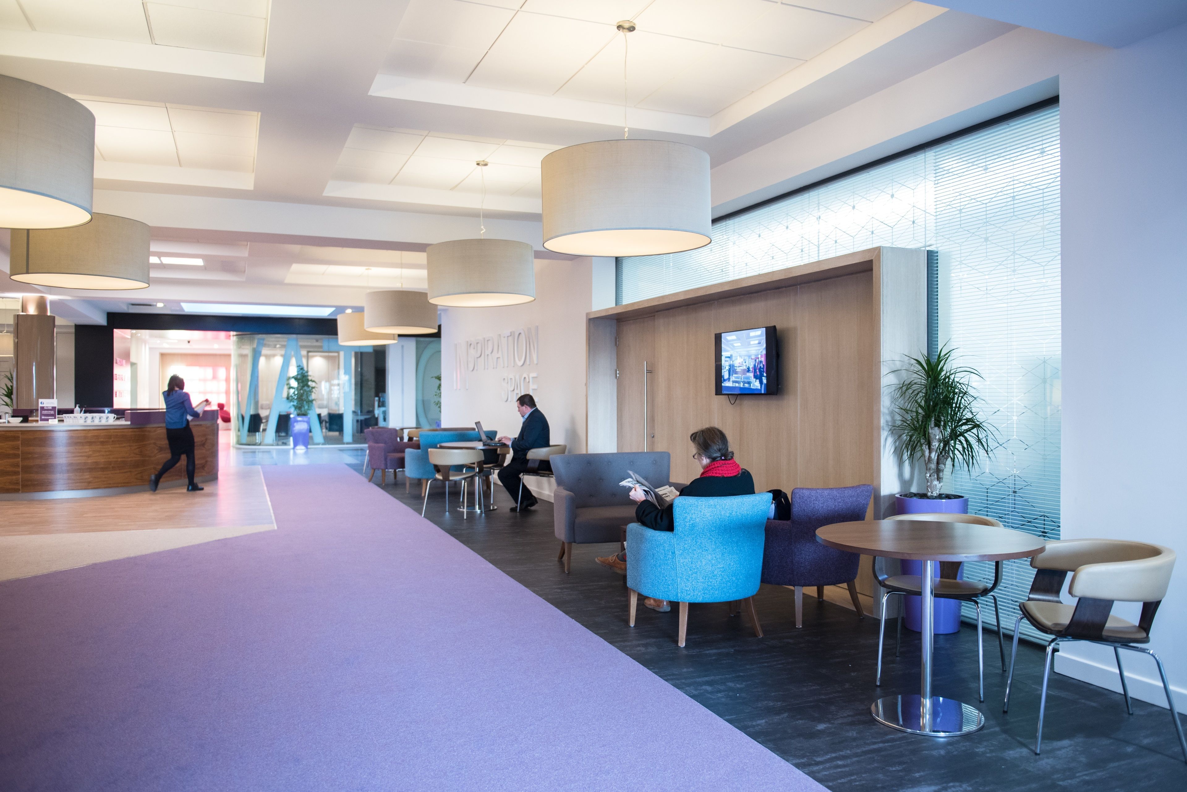 A modern lobby with a reception desk, seating areas, and a large window with blinds.