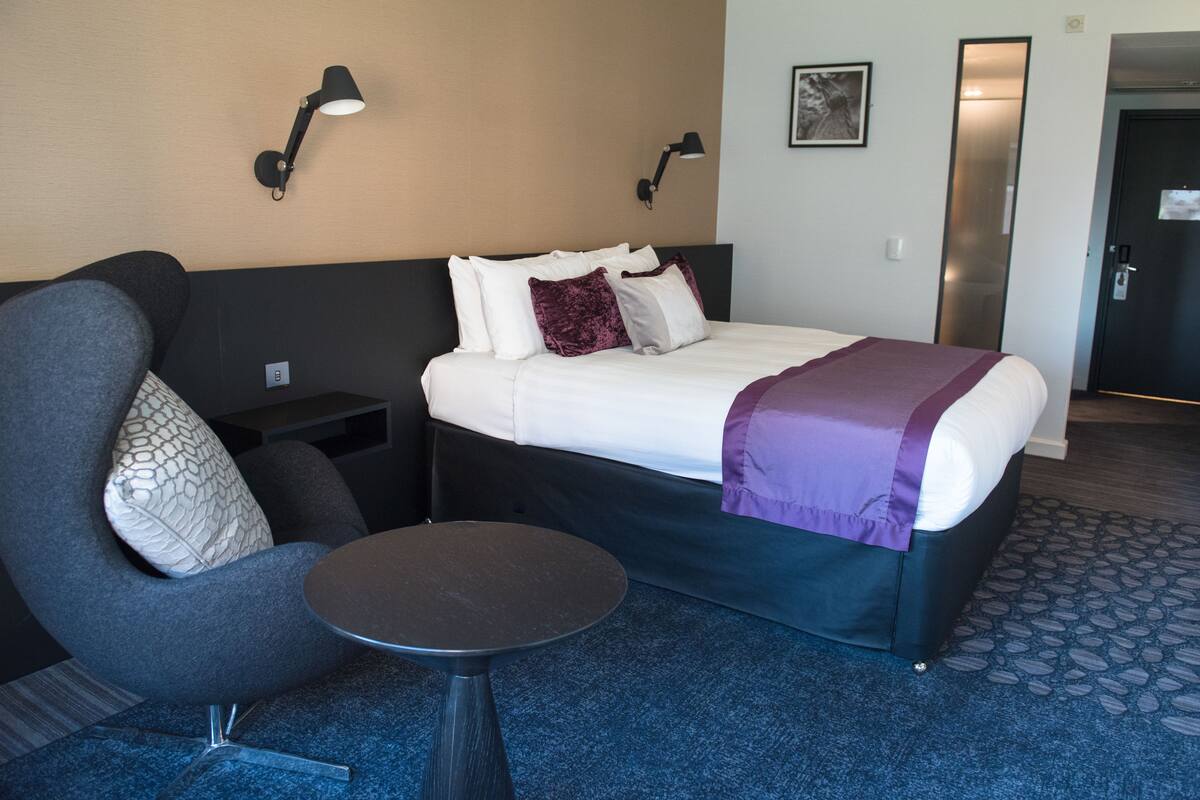 Una habitación de hotel moderna con una cama, una silla, una mesita y dos lámparas fijadas a la pared.