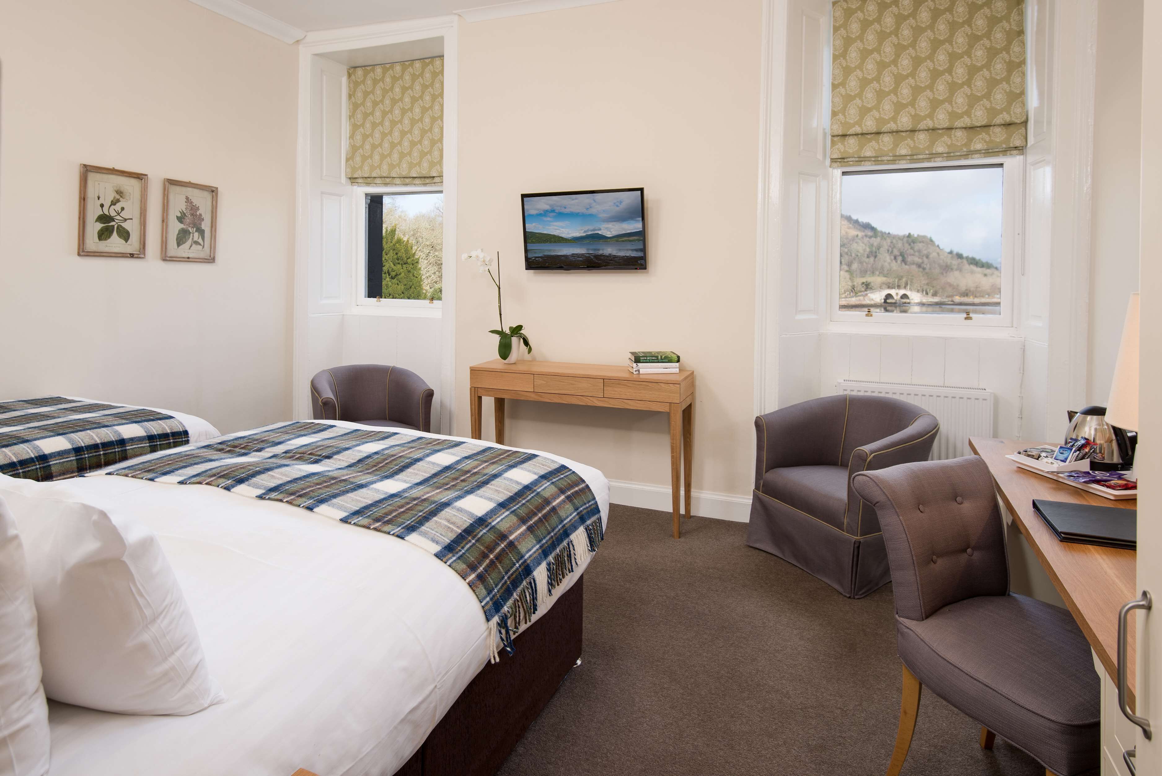 A hotel room with two beds, a desk, a chair, and a window with a view of a bridge and hills.
