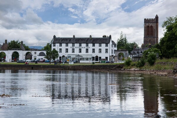 A white building with "The Inveraray Inn" sign, located beside a body of water with a tower in the background.