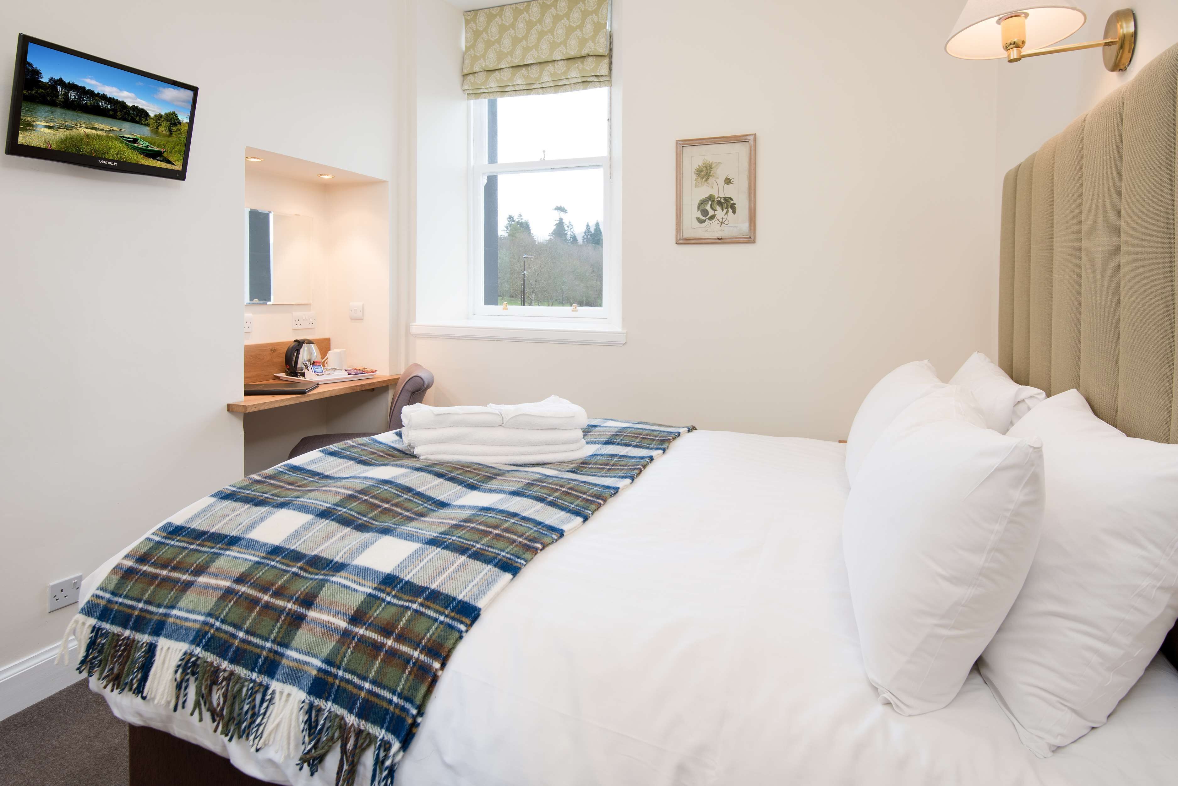 A bedroom with a bed, a bedside table, a window, a wall-mounted TV, and a framed picture.
