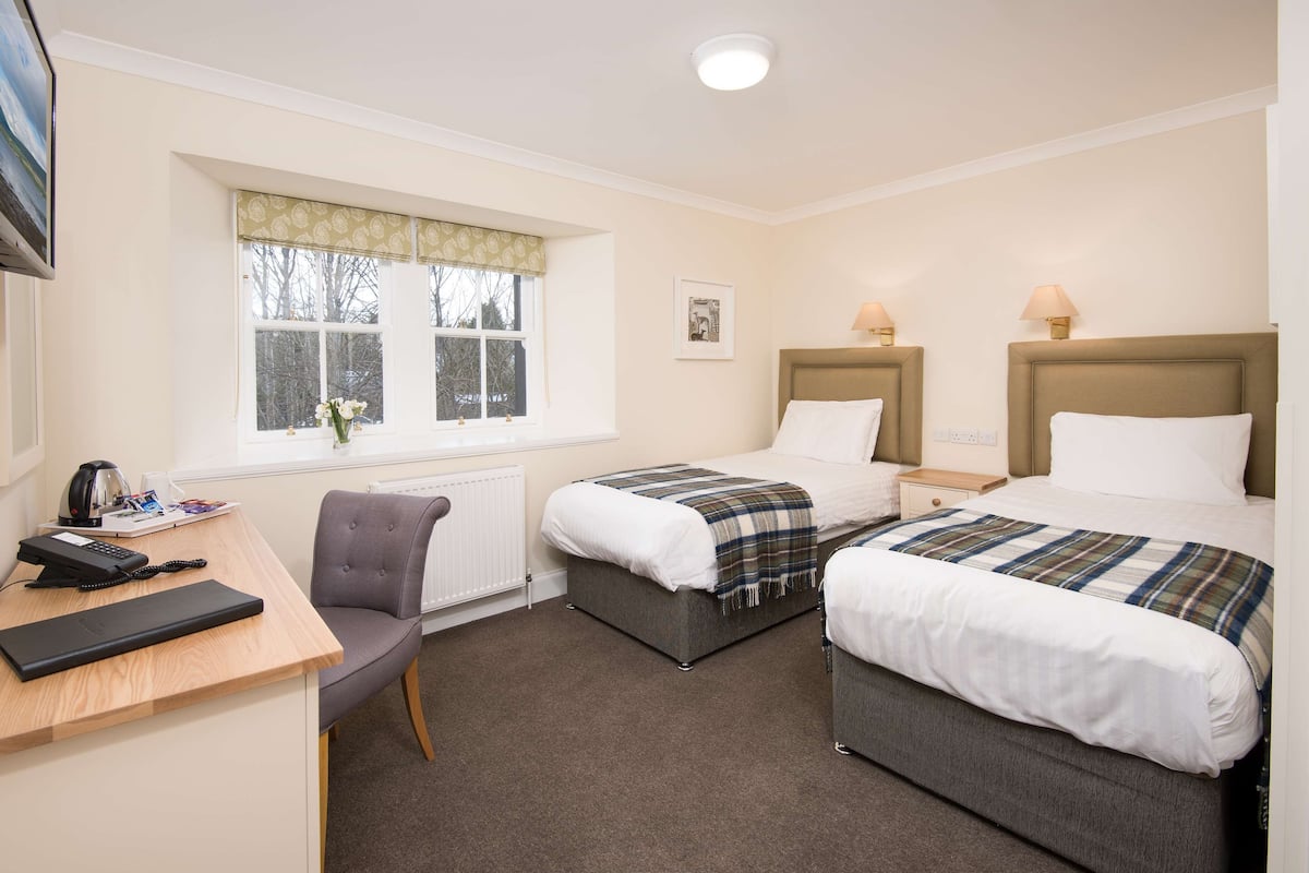 A hotel room with two single beds, a desk with a telephone, a chair, and a window with a view of trees.