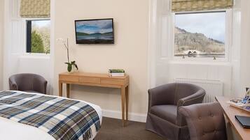 A hotel room with a bed, a desk, a chair, a TV, and a window with a view of a lake and hills.