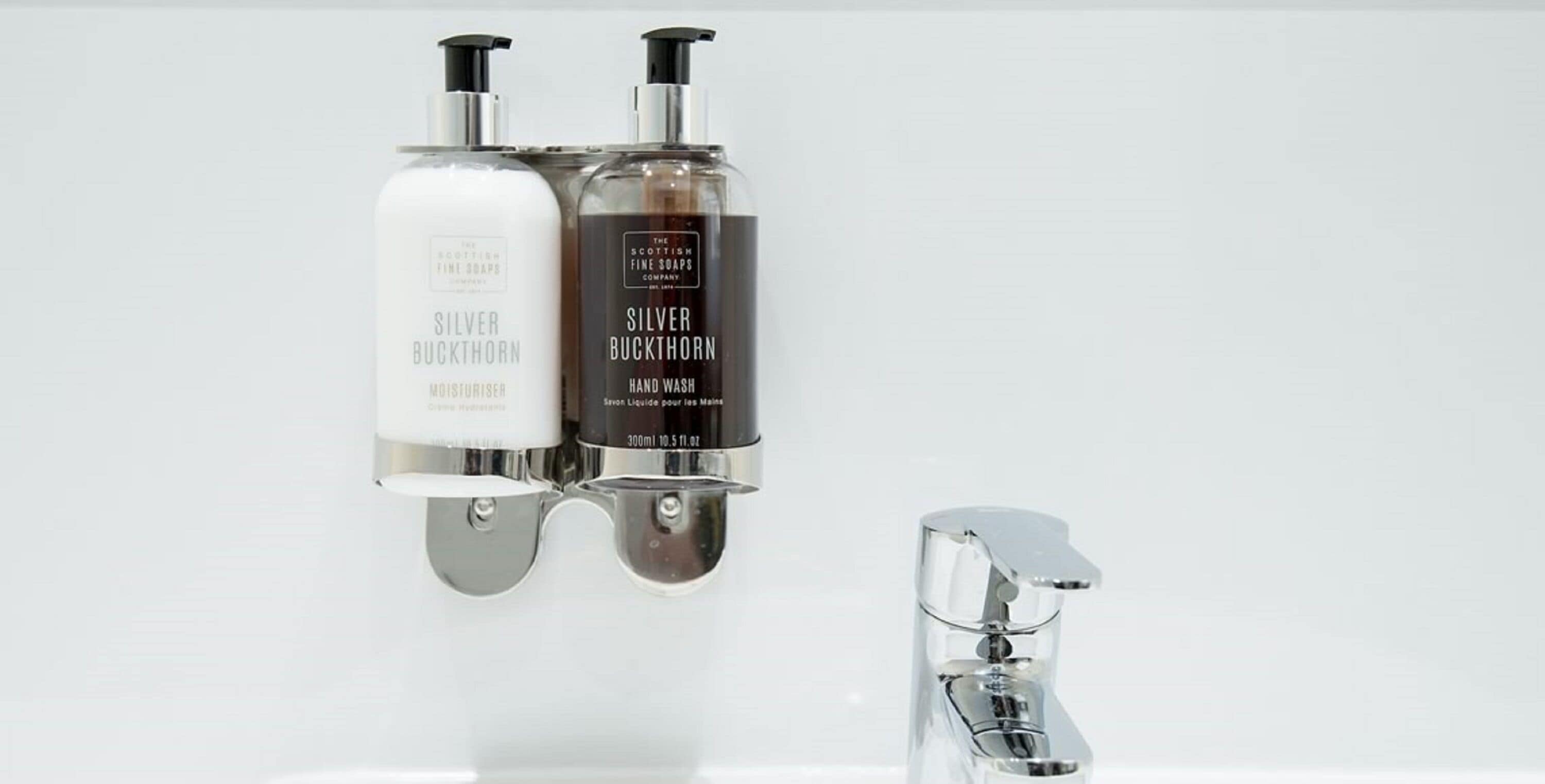 Two bottles of Silver Buckthorn hand wash soap mounted on a wall next to a chrome faucet.