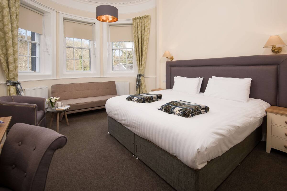 A neatly arranged bedroom with a large bed, a sofa, a small table with a lamp, and a window with curtains.