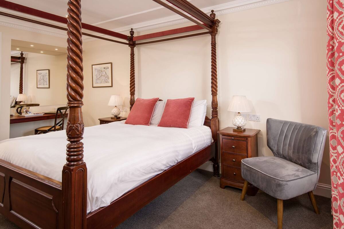 A four-poster bed with a wooden frame, a grey upholstered armchair, and a wooden nightstand with a lamp.