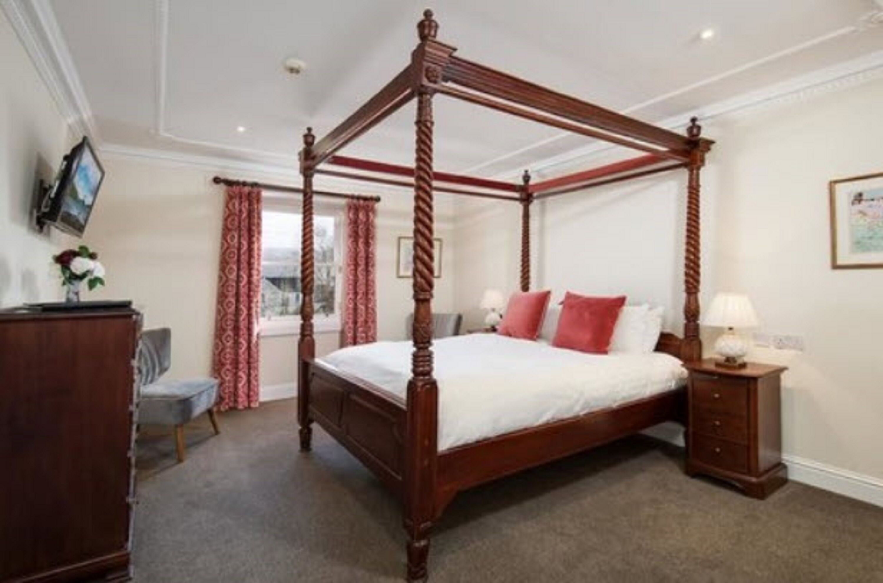 A four-poster bed with a wooden frame, a bedside table with a lamp, and a chair in a room with a carpeted floor and a window with curtains.