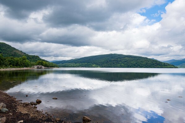 Un lac paisible, avec un pont qui se dessine au loin, entouré de collines verdoyantes et surplombé d’un ciel nuageux.