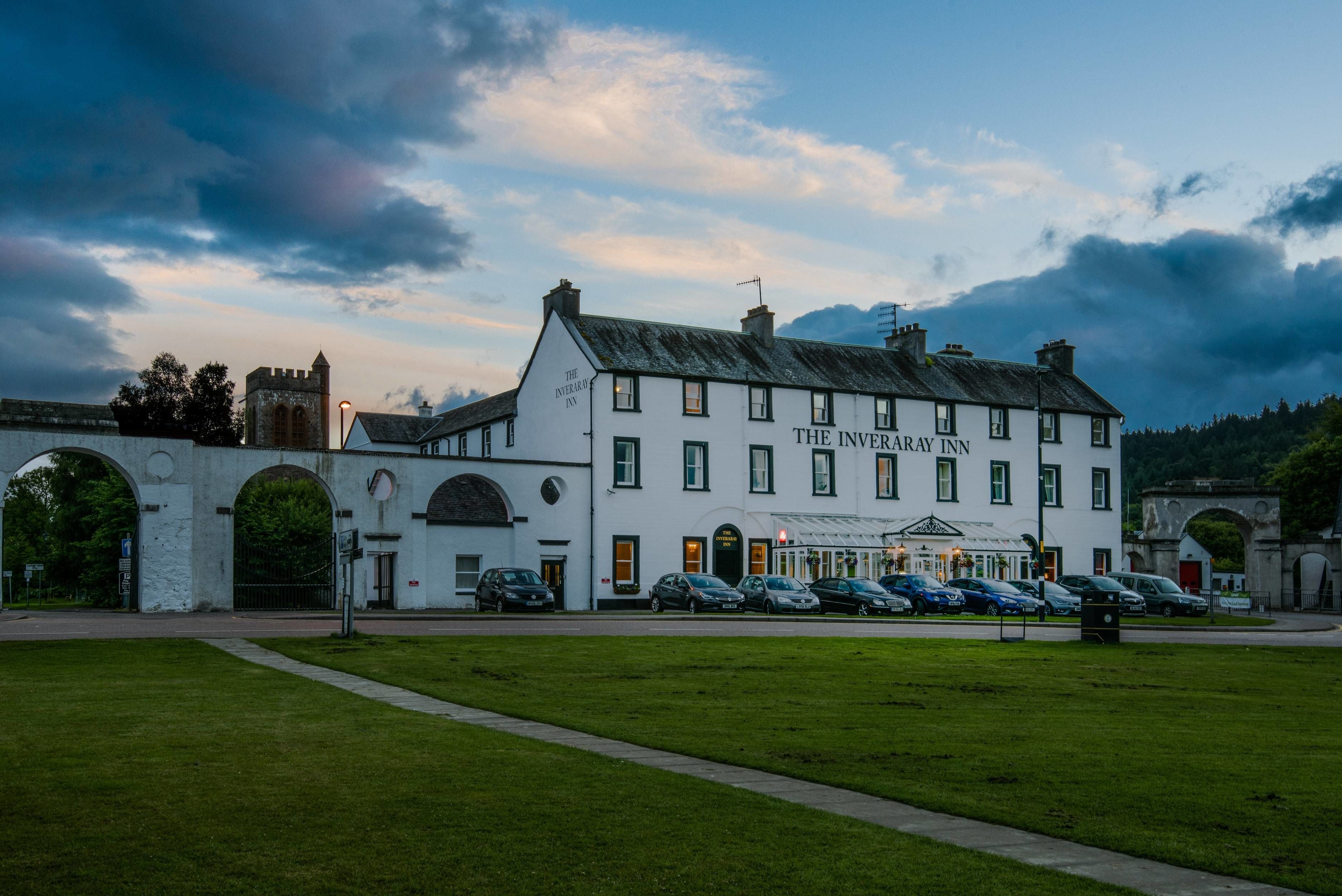 L’hôtel Inveraray, un bâtiment blanc doté de nombreuses fenêtres et d’une enseigne, est situé dans une zone verdoyante, avec des voitures garées et une structure en arc de cercle en arrière-plan.