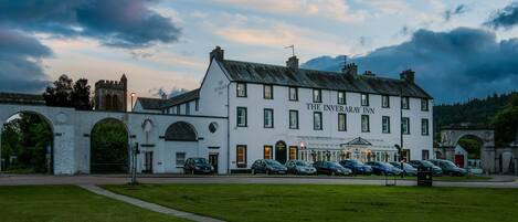 L’hôtel Inveraray, un bâtiment blanc doté de nombreuses fenêtres et d’une enseigne, est situé dans une zone verdoyante, avec des voitures garées et une structure en arc de cercle en arrière-plan.