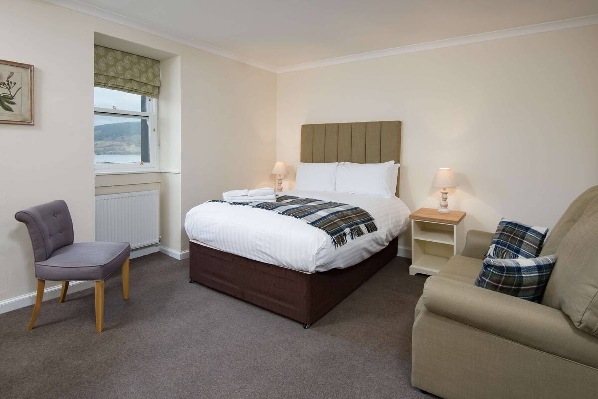 A bedroom with a bed, two bedside tables with lamps, a chair, and a view of the outdoors through a window.