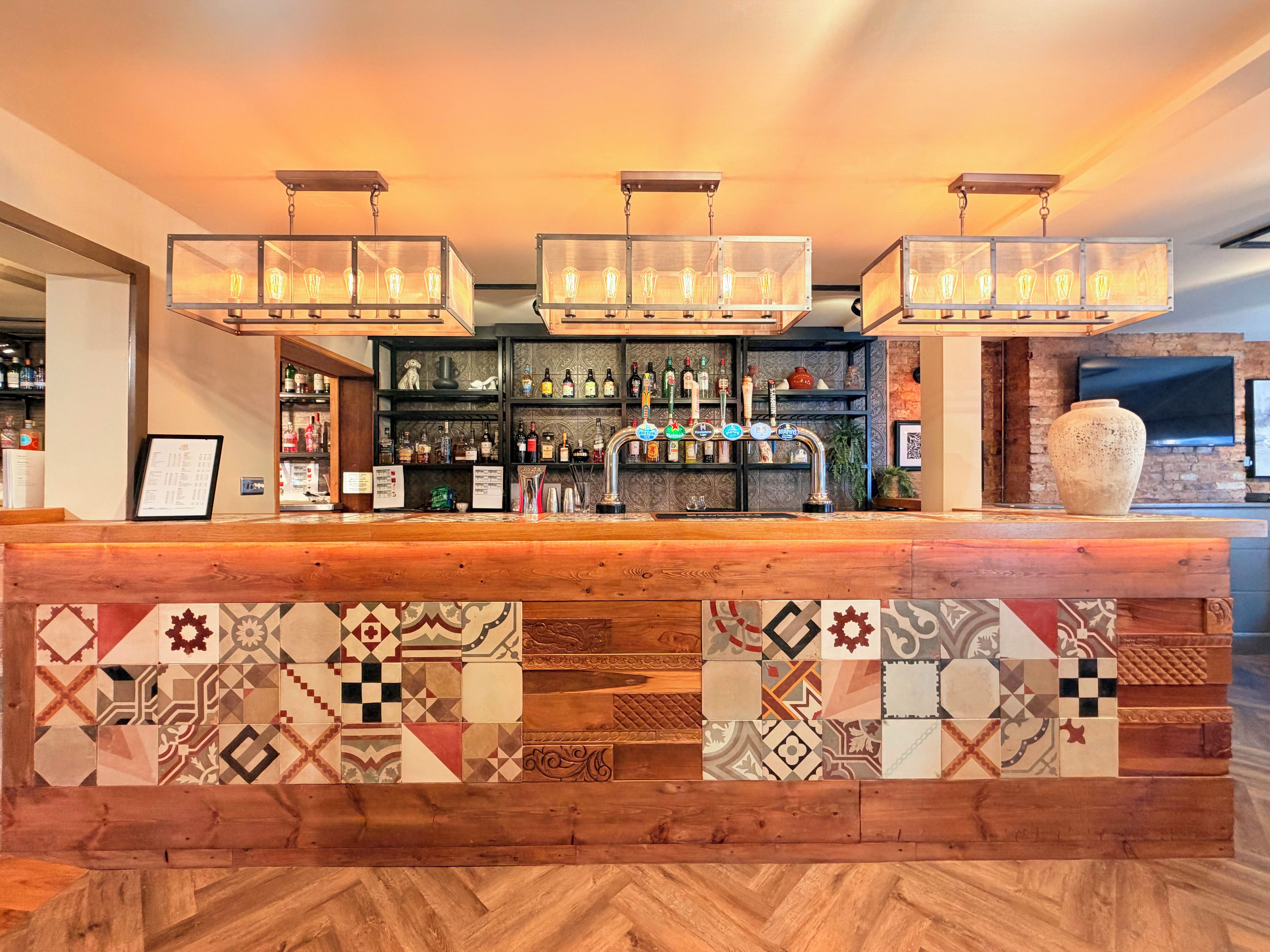 A bar area with a wooden counter, multiple light fixtures, and a variety of bottles and glasses.