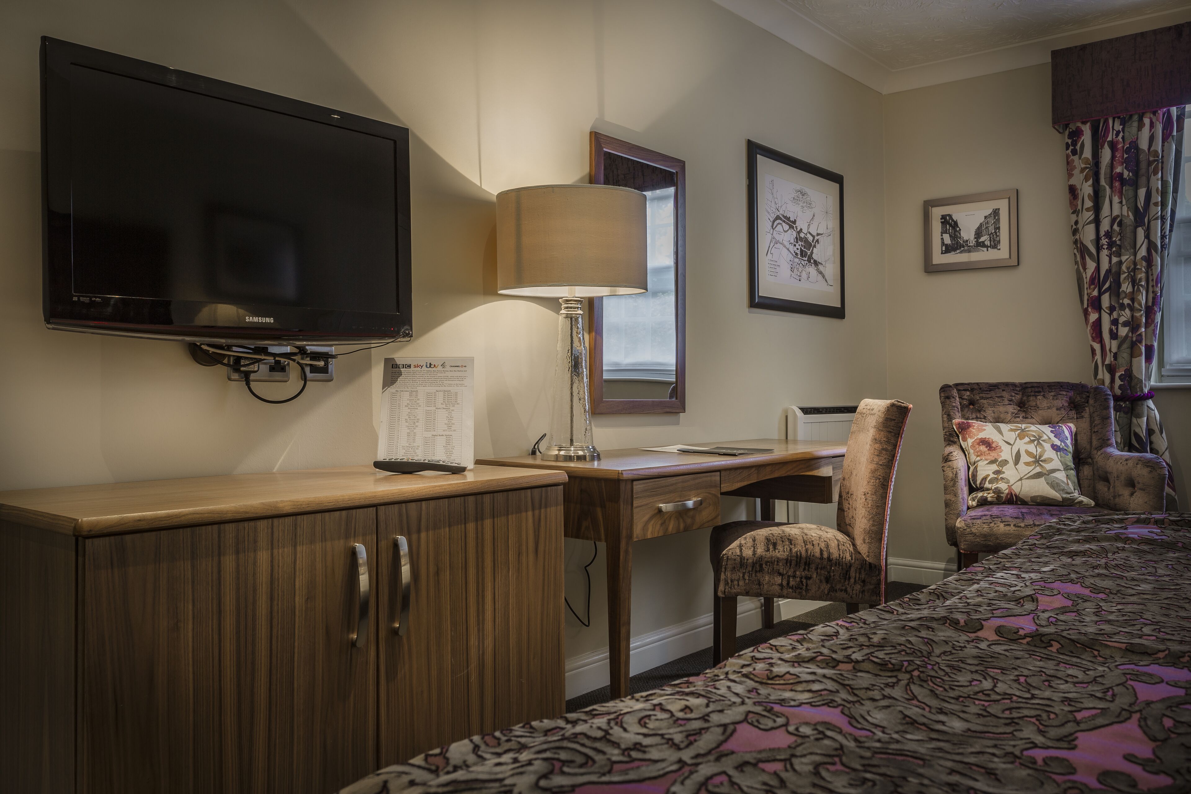 Habitación de hotel con televisor de pantalla plana, un escritorio con lámpara, una silla, cama con colcha estampada y cuadros en la pared.