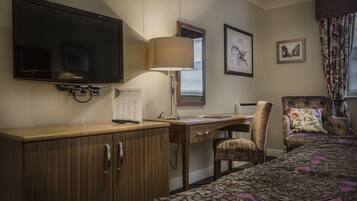 A hotel room with a flat-screen TV, a desk with a lamp, a chair, a bed with a patterned bedspread, and framed pictures on the wall.