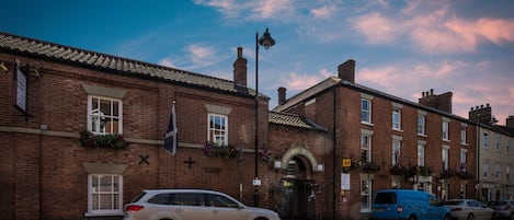 A brick building with a traditional arched entrance, a blue van parked in front, and a beige car parked on the street.