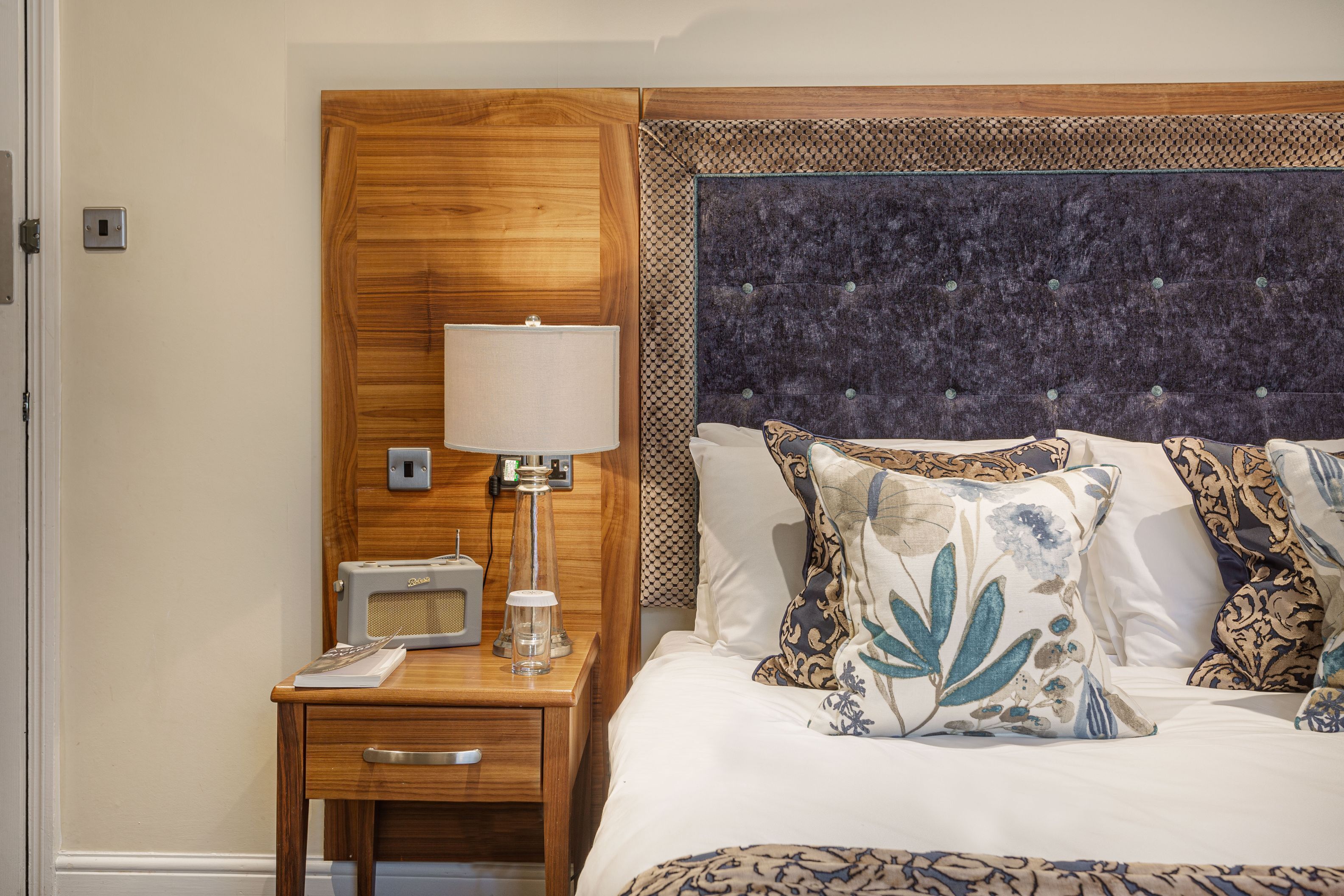 Une chambre d’hôtel avec une tête de lit en bois, une table de chevet avec une lampe, une radio et des coussins décoratifs.