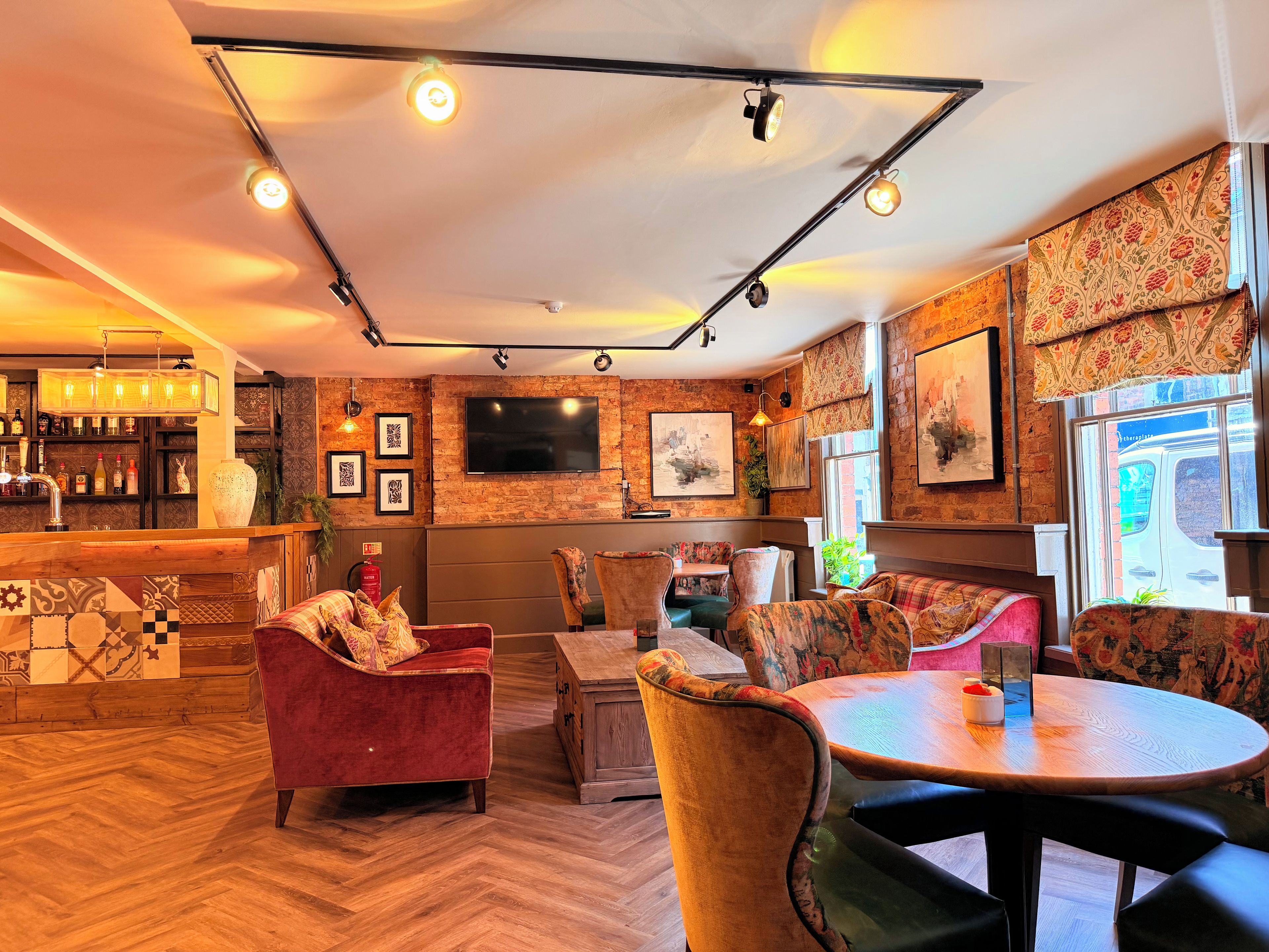 A cozy bar area with a wooden counter, plush seating, and a large TV mounted on a brick wall.
