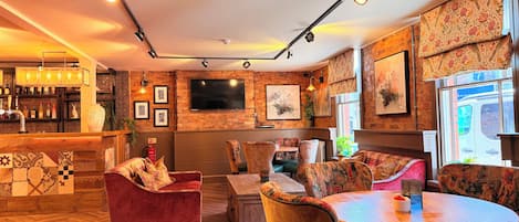 A cozy bar area with a wooden counter, plush seating, and a large TV mounted on a brick wall.