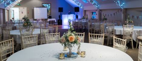A banquet hall with round tables, chairs, and a central floral arrangement.