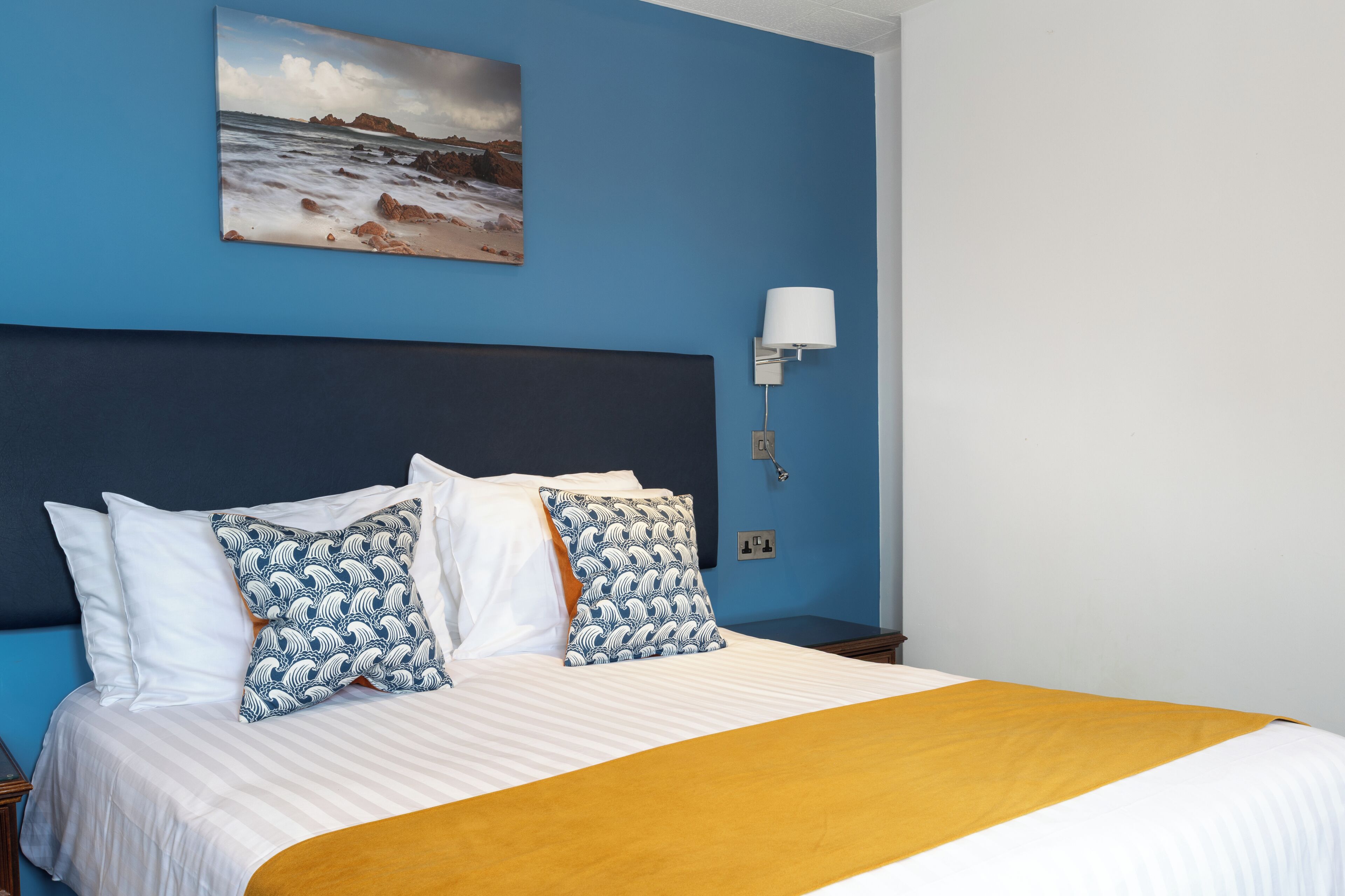 Una cama con almohadas de diseño blanco y azul, una colcha a rayas y una pared azul adornada con un cuadro de playa.