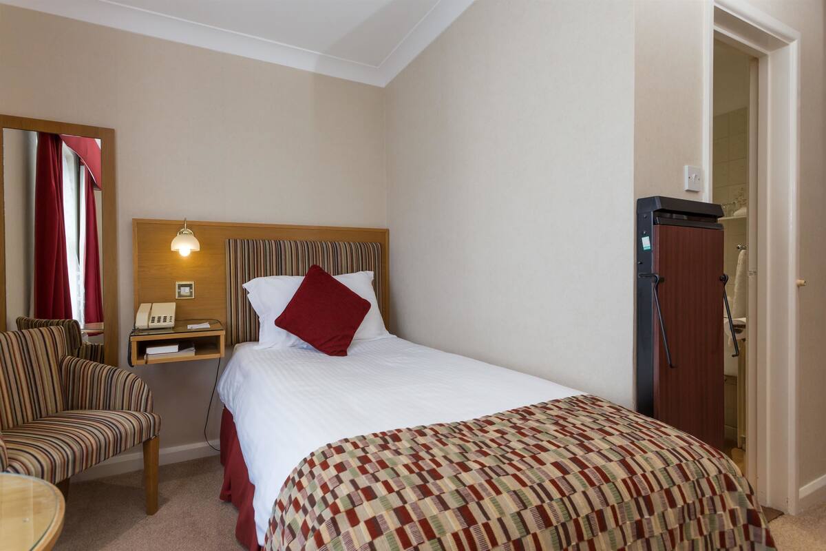Une chambre d’hôtel comprenant un lit, une chaise, une table de chevet avec une lampe et une porte donnant accès à une salle de bain.