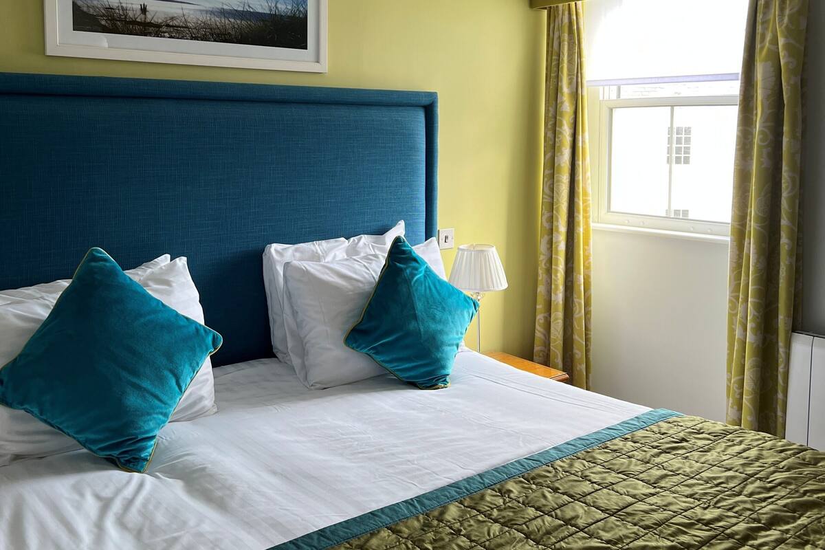 Une chambre d’hôtel avec un lit, une tête de lit, un tableau encadré au mur, une fenêtre avec des rideaux et une lampe de chevet.