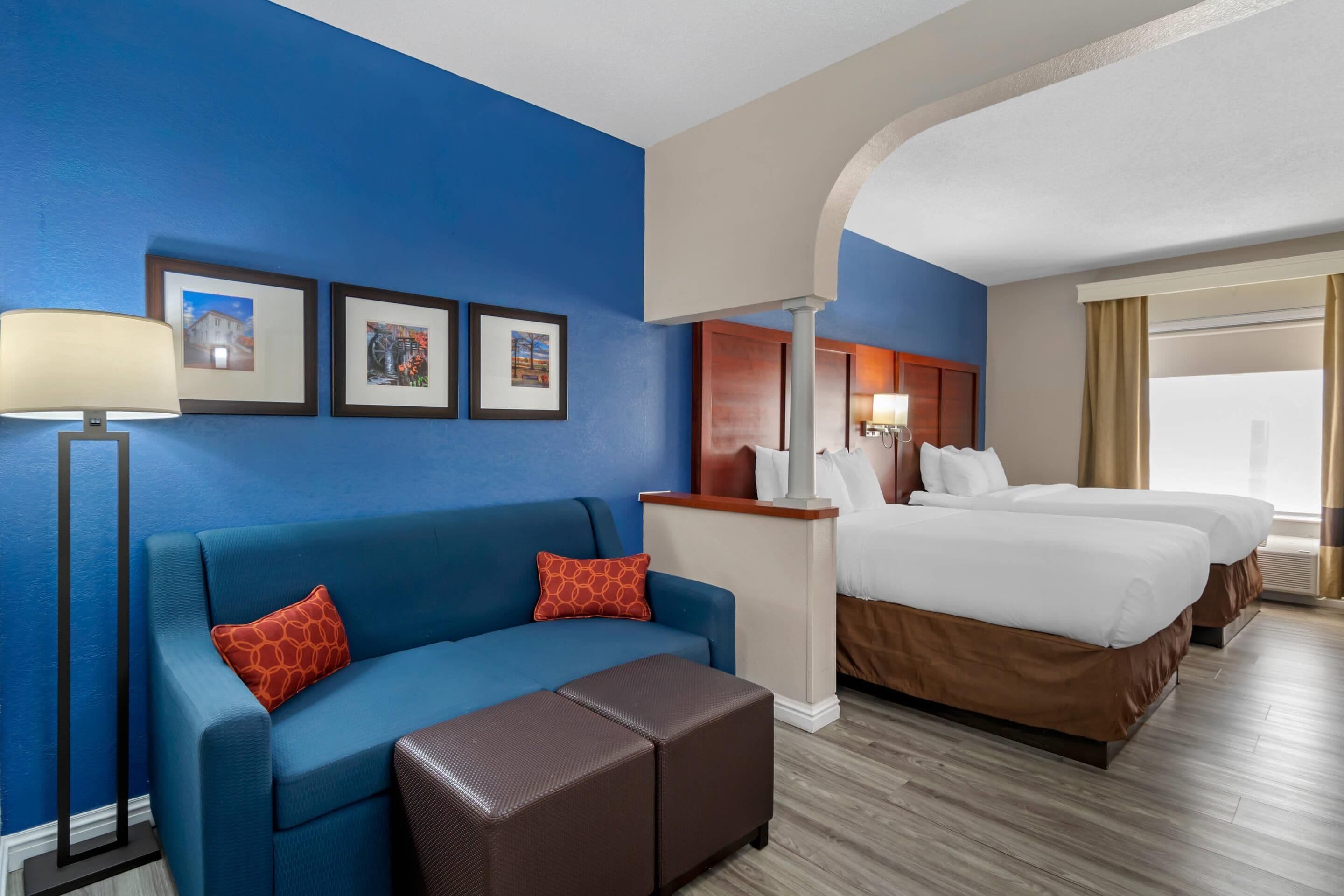 Quarto de hotel com parede azul, sofá, banqueta marrom, cama com roupa de cama branca e mesa de cabeceira com abajur.