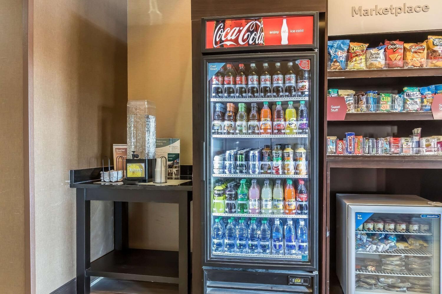 A vending machine with Coca-Cola and various beverages, a small table with a water dispenser, and shelves with snacks and ice boxes.