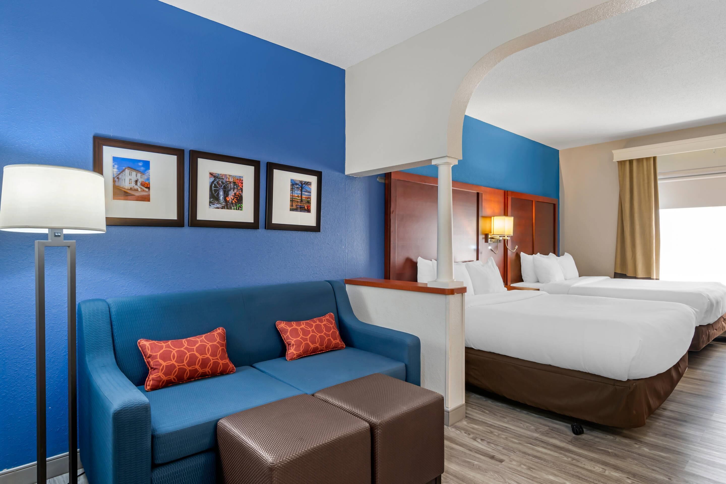 Ein Hotelzimmer mit blauer Wand, einer Couch mit roten Kissen in Musteroptik, einem braunen Hocker, einem Bett mit weißen Bettwäsche und gerahmten Bildern an der Wand.
