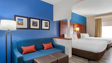 Ein Hotelzimmer mit blauer Wand, einer Couch mit roten Kissen in Musteroptik, einem braunen Hocker, einem Bett mit weißen Bettwäsche und gerahmten Bildern an der Wand.
