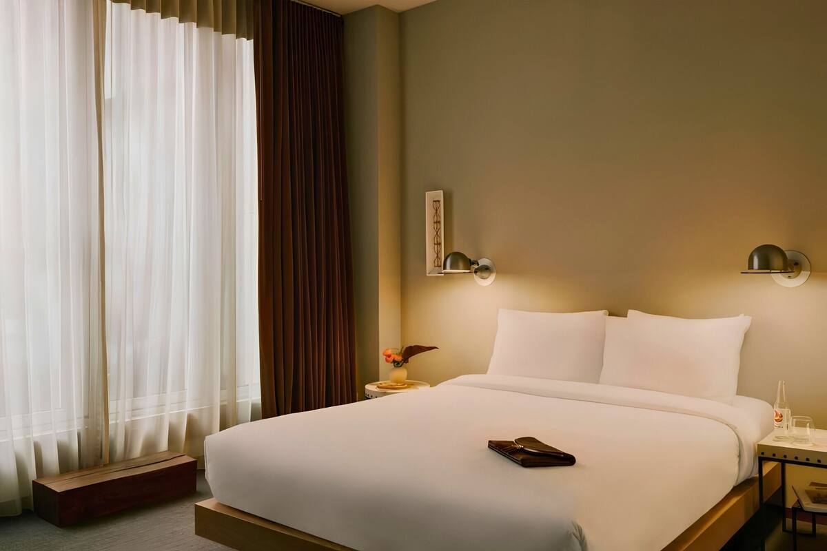 Habitación de hotel con cama, mesita de noche, lámpara de pared y ventana con cortinas.
