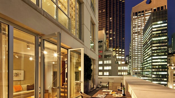 Una habitación de hotel moderna con un balcón accesible mediante una puerta de vidrio, un área de comedor con mesa y sillas, y vistas de edificios altos de noche.