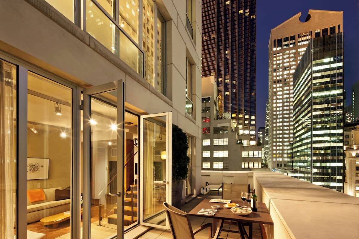Una habitación de hotel moderna con un balcón accesible mediante una puerta de vidrio, un área de comedor con mesa y sillas, y vistas de edificios altos de noche.