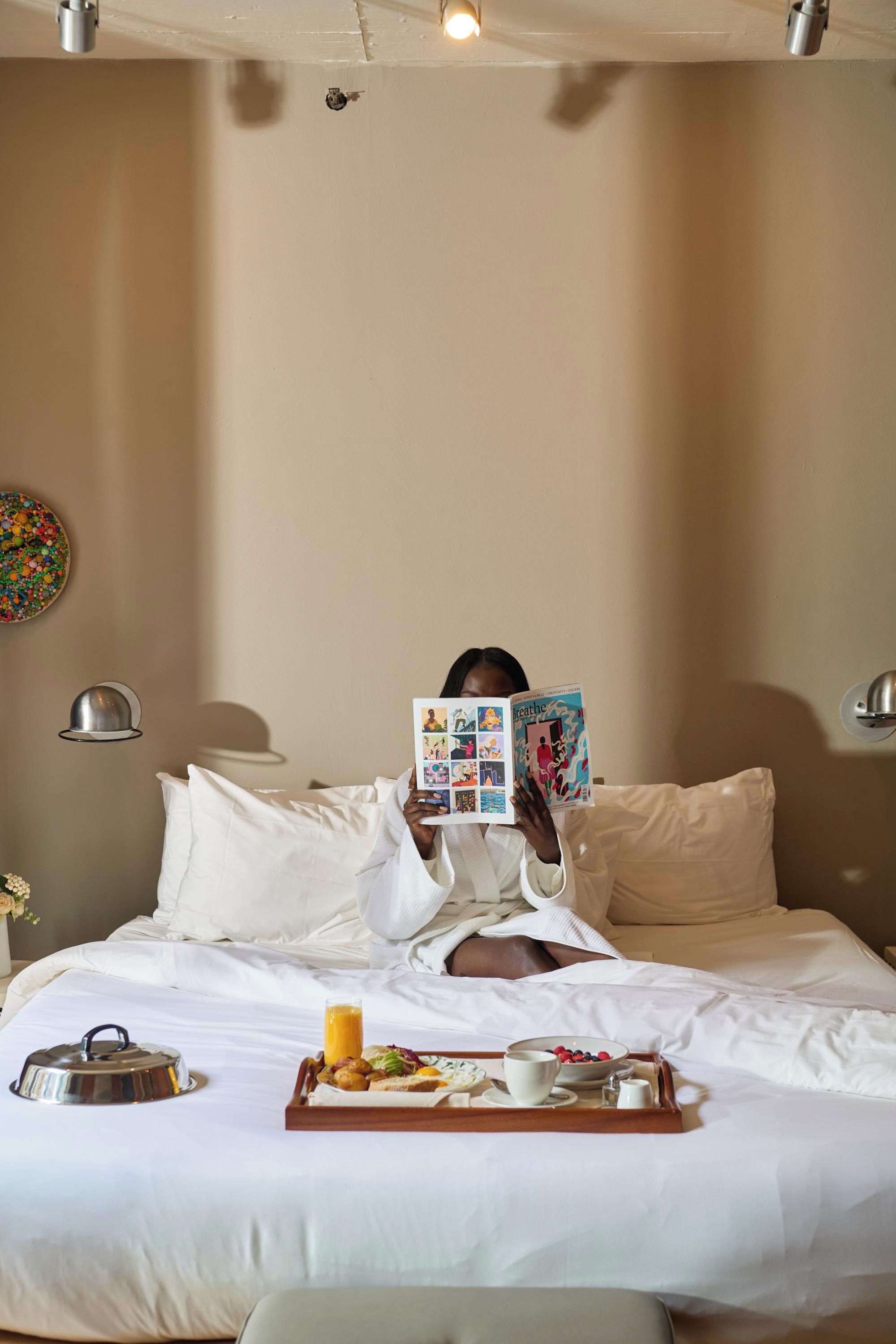 A person lying in bed reading a magazine, with a tray of breakfast items beside them.