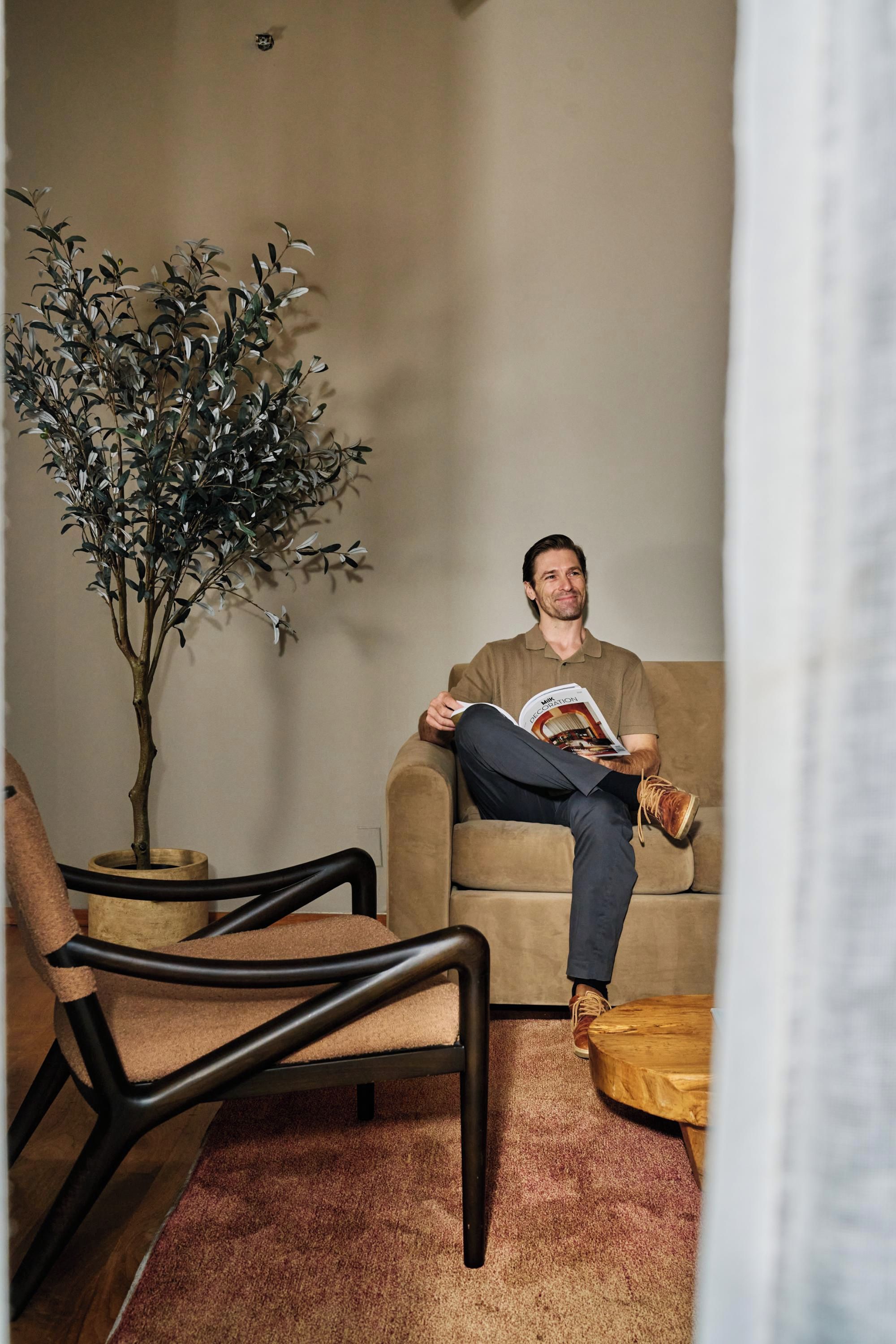 Un homme est assis sur un canapé, avec un magazine, une plante en pot et une chaise, dans une pièce dont le sol est recouvert d’une moquette.