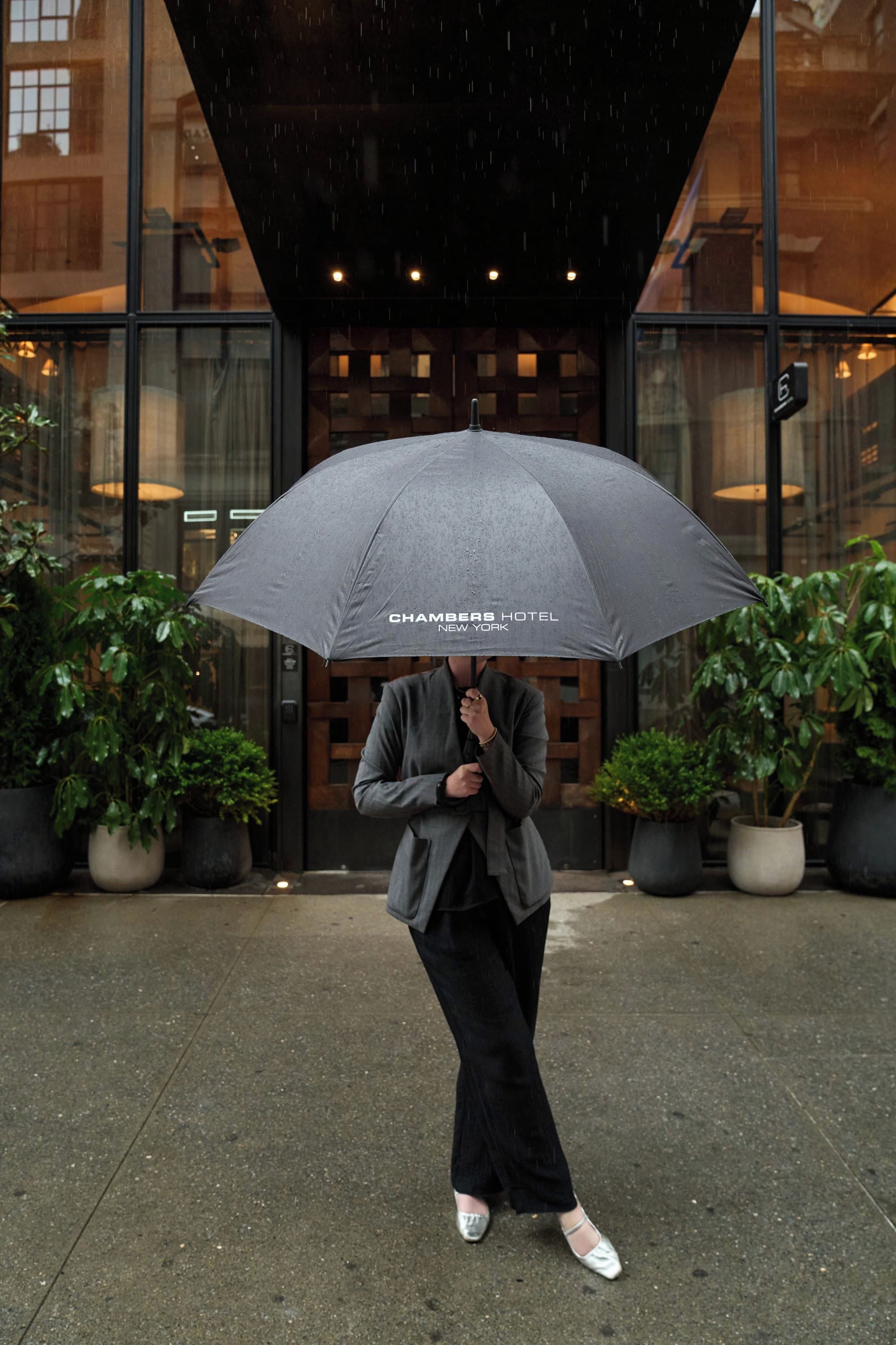 A person standing under an umbrella in front of a building with a glass facade and potted plants.